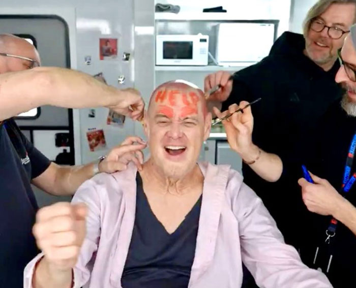 Man laughing as three people paint unhinged behind-the-scenes photos on his bald head in a playful setting.