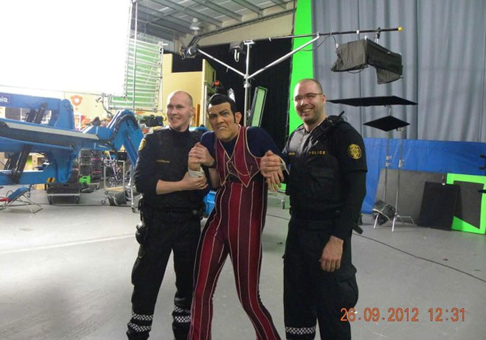 Behind-the-scenes photo of two men in police uniforms holding a man in a striped costume on a film set with green screen.