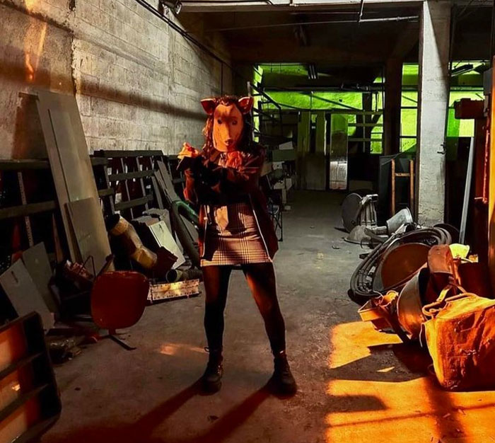 Person wearing an unhinged animal mask posing in a cluttered behind-the-scenes room with dramatic lighting and shadows.