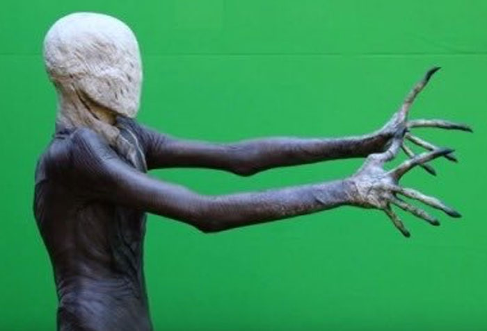 Unhinged behind-the-scenes photo of a creature with elongated arms and clawed hands in front of a green screen.