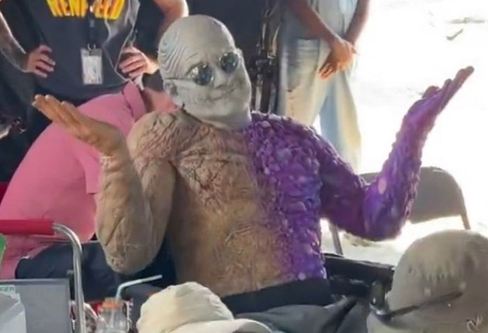 Actor in unhinged behind-the-scenes costume with half purple, half beige textured makeup shrugging on set during a casual moment.