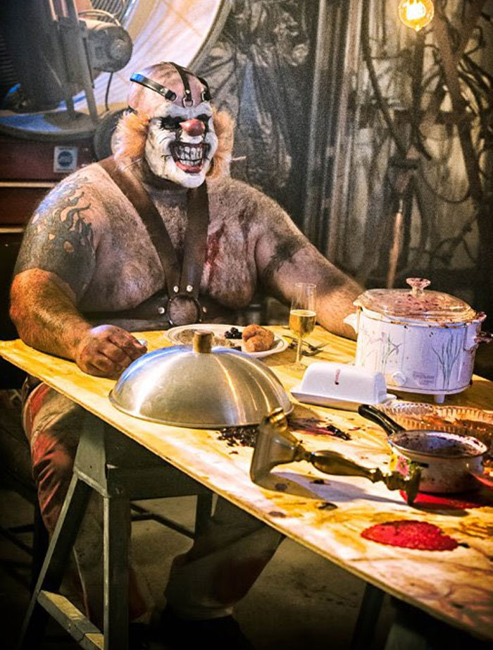 Unhinged behind-the-scenes photo of a muscular man in clown makeup sitting at a messy table with food props.