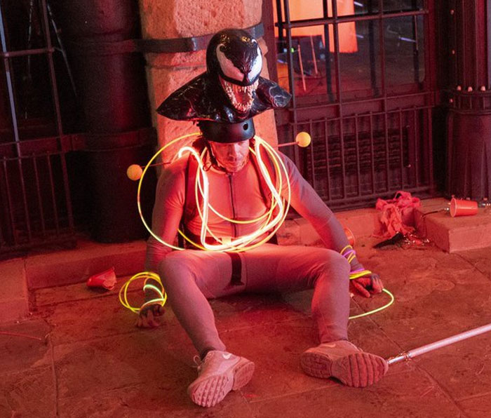Man in a Venom helmet and light-up wires sitting unhinged on the ground in a behind-the-scenes photo meme moment.