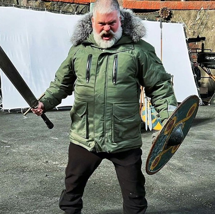 Man in a green parka brandishing a sword and shield on set, one of the unhinged behind-the-scenes photos meme moments.