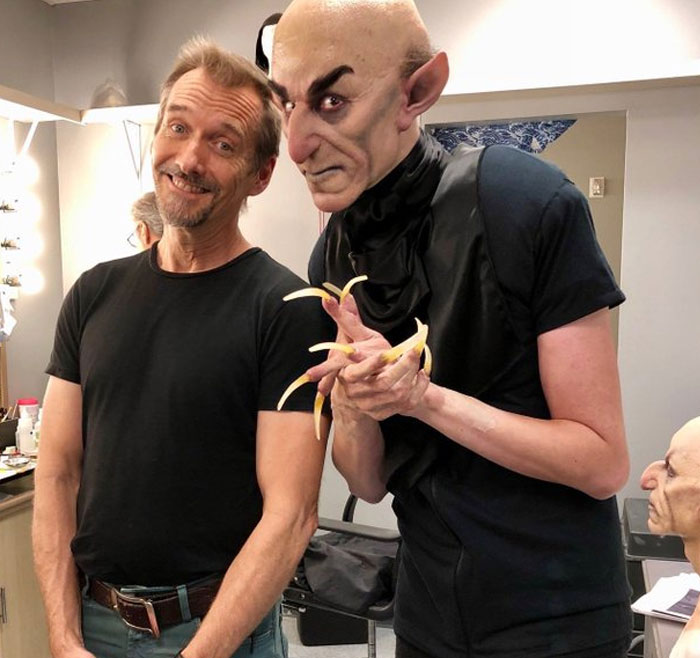 Behind-the-scenes photo of actor with makeup artist in unusual costume and long claw-like nails on set.