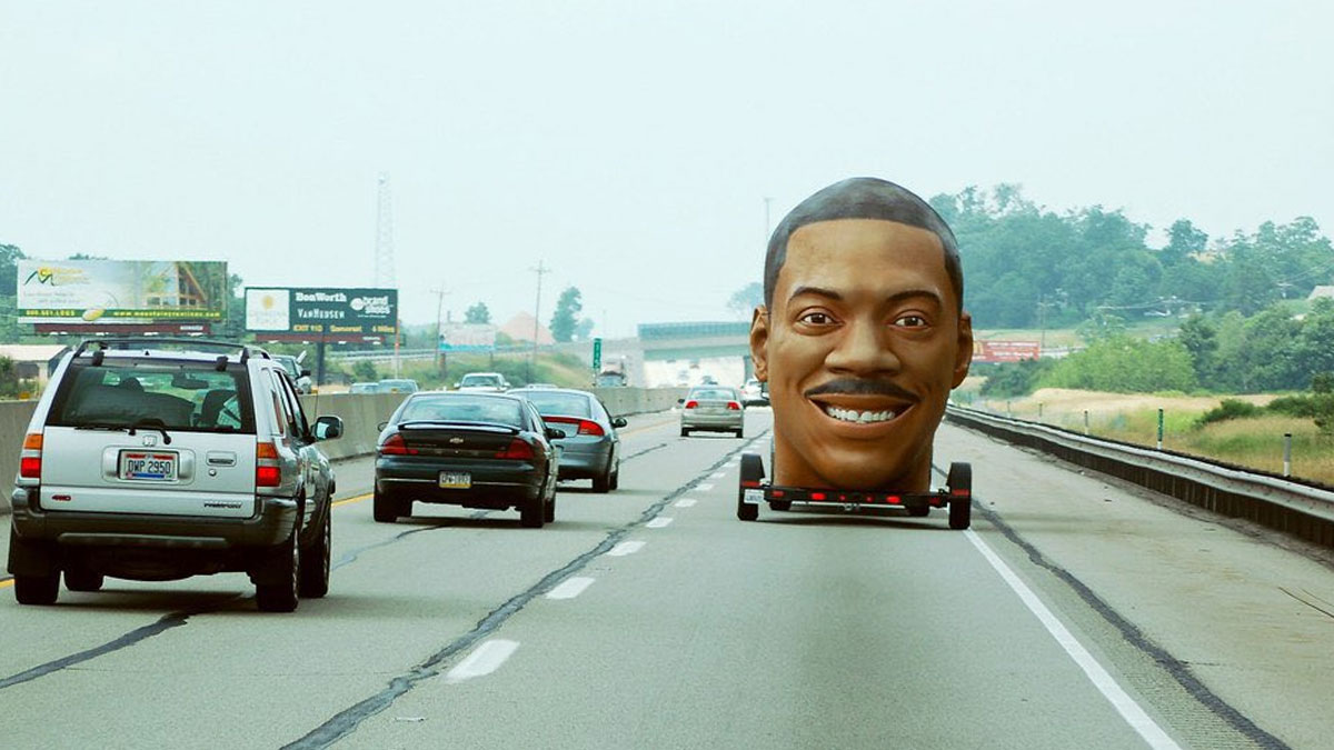 Unhinged behind-the-scenes photo of a huge smiling head on a trailer driving on a highway among regular cars.