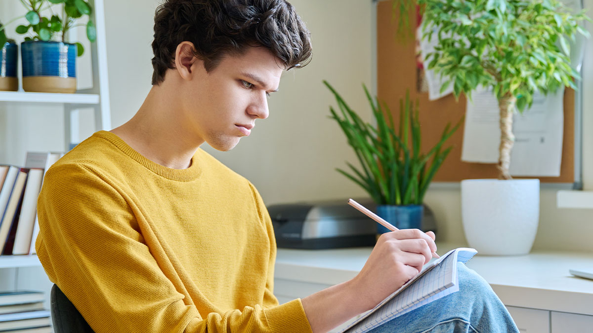 Teen writing a note indoors, deeply focused, symbolizing a reality check gift on his father's 50th birthday celebration.