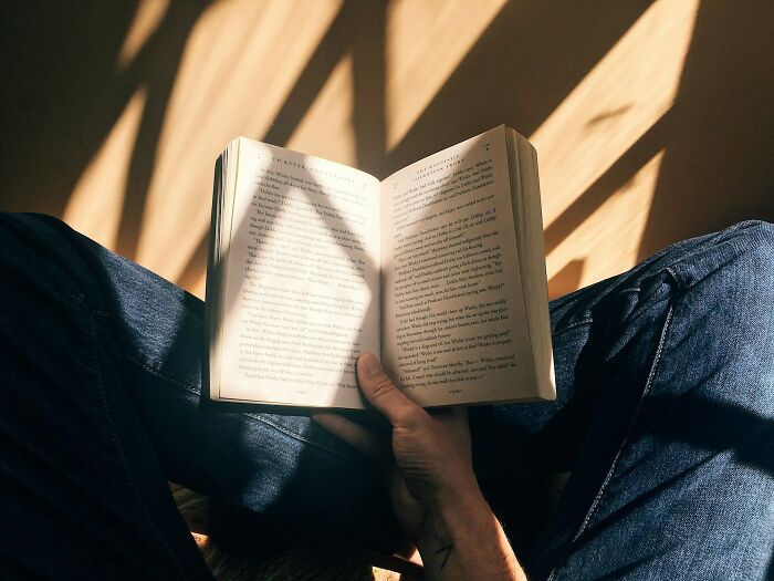 Person reading a book on etymology of common words with sunlight casting shadows on the pages and jeans.