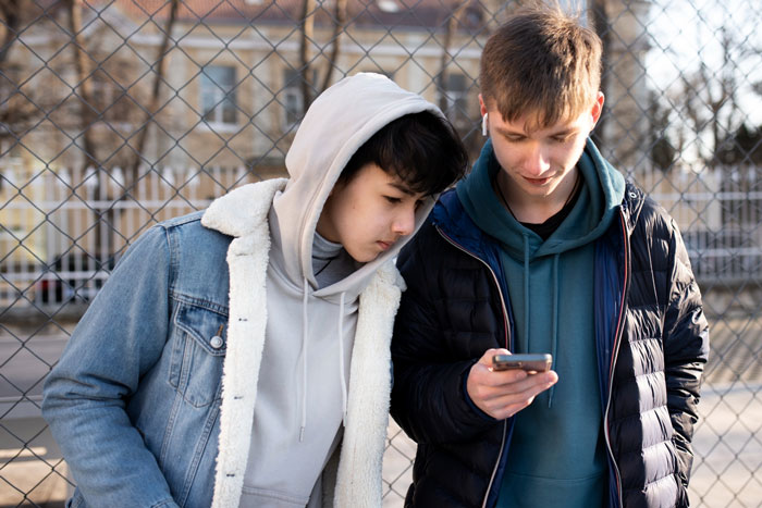 Two teenage boys outdoors near a fence, one laughing while showing something on a smartphone, capturing sibling rivalry tension. Two teenage boys outdoors near a fence, one laughing while showing something on a smartphone, capturing sibling rivalry tension.