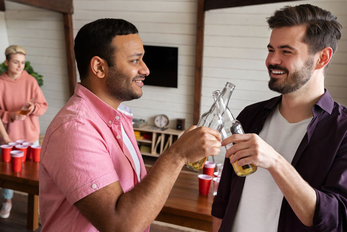 Two men clinking beer bottles at a casual party, while a woman stands in the background near a table with red cups. Two men clinking beer bottles at a casual party, while a woman stands in the background near a table with red cups.