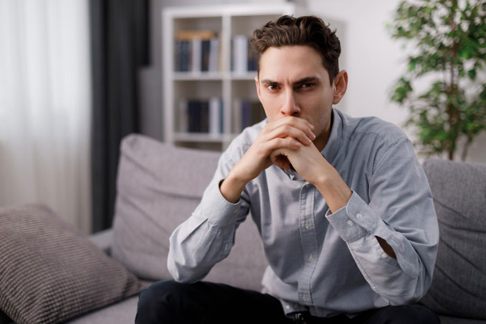 Young worried man sitting on couch with hands clasped, reflecting on relationship trust and concerns about cheating.