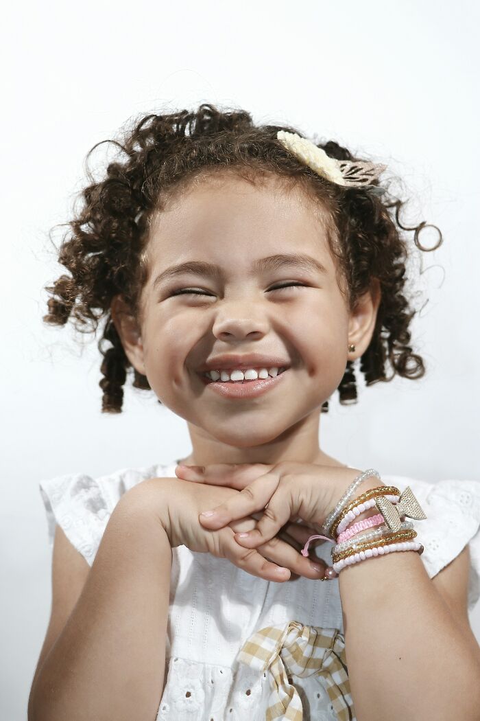 Smiling curly-haired girl wearing bracelets and a white dress, capturing funny and serious moments of raising children.