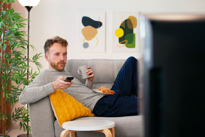 Man relaxing on couch watching TV with coffee and croissants, illustrating nanny with benefits relationship concept.