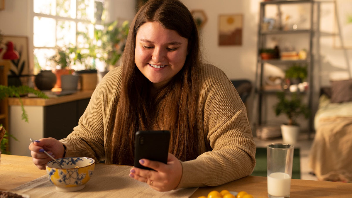 Young woman smiling while looking at phone, eating breakfast, illustrating sibling mocking and diabetes theme.