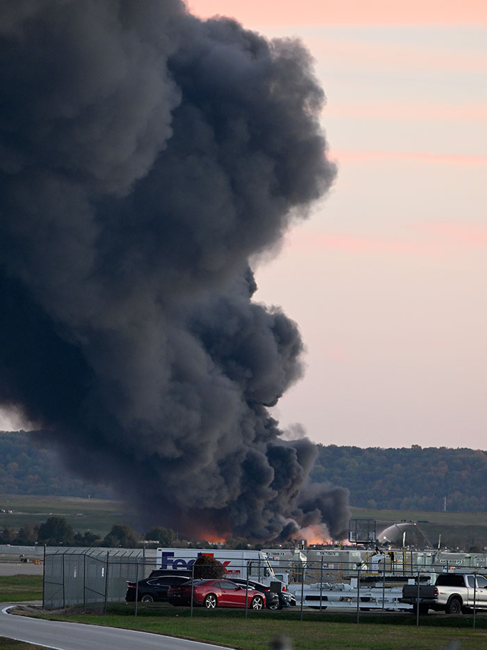 Thick black smoke billowing from a UPS plane crash site with emergency response vehicles nearby at sunset. Thick black smoke billowing from a UPS plane crash site with emergency response vehicles nearby at sunset.