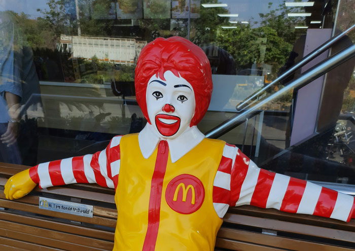 Ronald McDonald statue with red hair, striped sleeves, and yellow vest sitting on a bench outside a McDonald's restaurant
