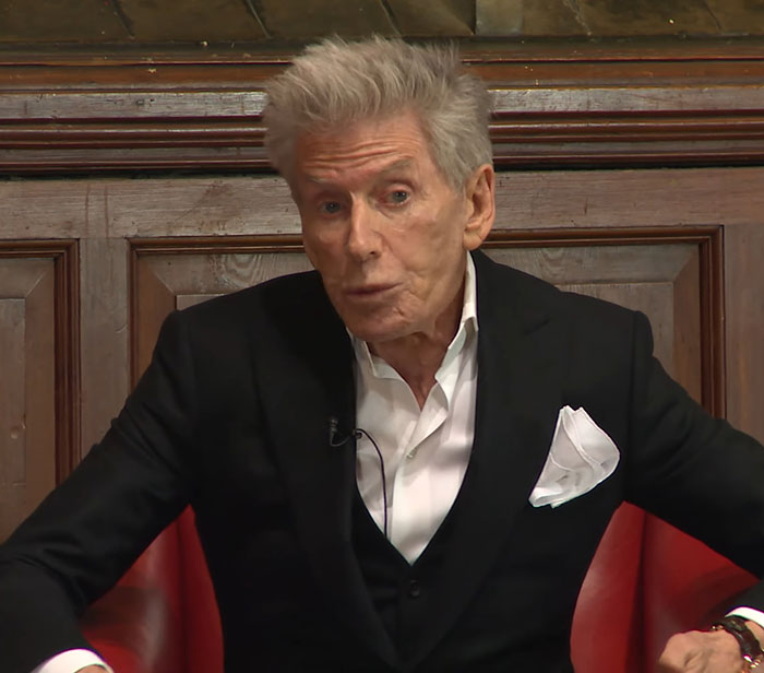 Calvin Klein, 83, wearing a black suit and white shirt, seated and speaking with a serious expression indoors. Calvin Klein, 83, wearing a black suit and white shirt, seated and speaking with a serious expression indoors.