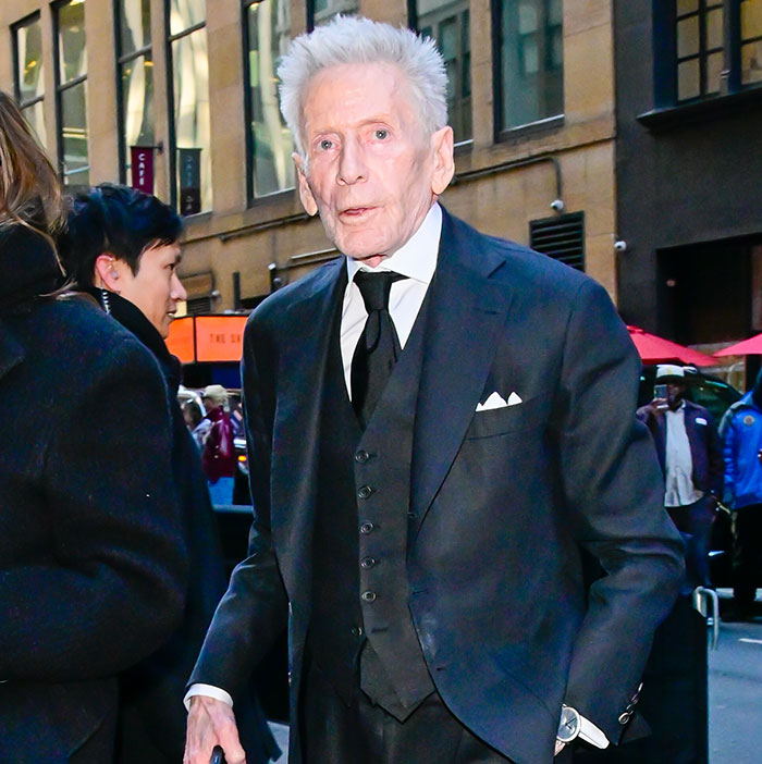 Calvin Klein, 83, wearing a black suit, walking outdoors with people around, capturing a candid moment in the city. Calvin Klein, 83, wearing a black suit, walking outdoors with people around, capturing a candid moment in the city.