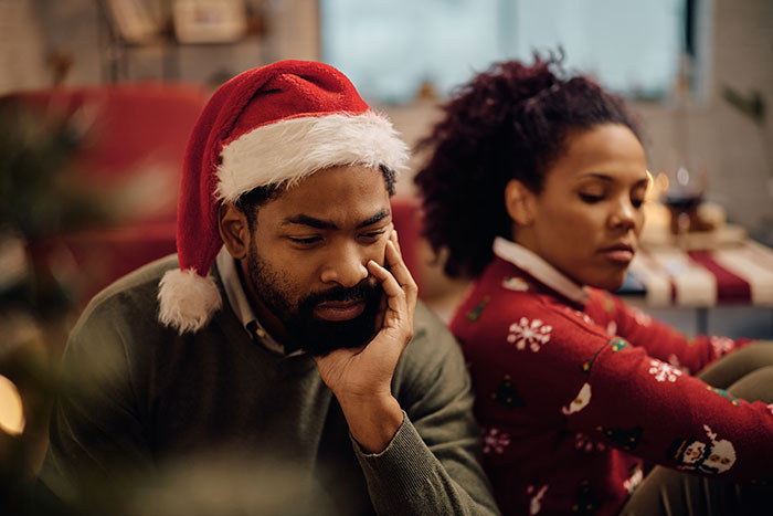 Father wearing Santa hat looks worried about Christmas expenses while woman in festive sweater sits nearby, both appearing upset.