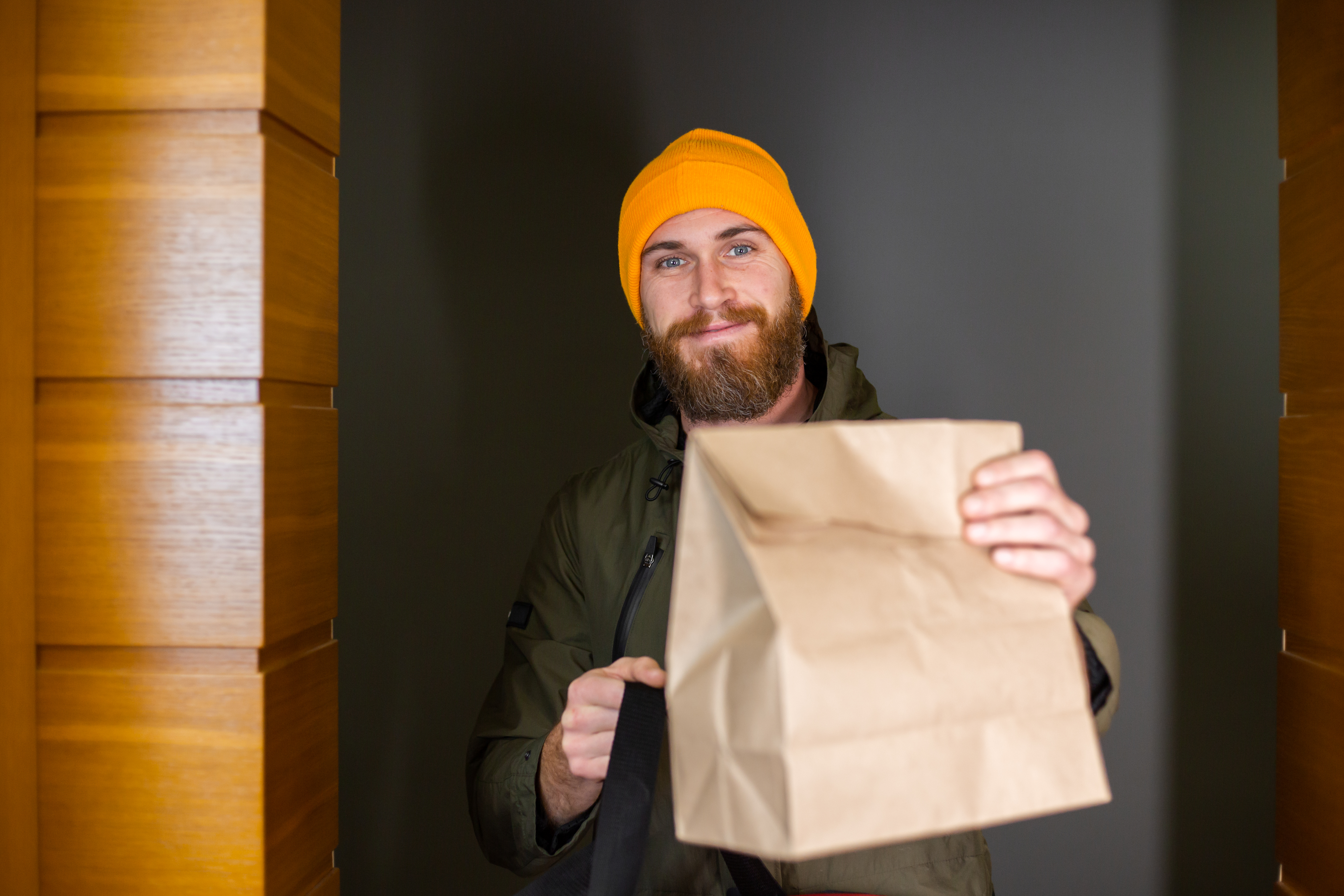 Smiling baker wearing a yellow beanie holding a paper bag for delivery, representing a bagel delivery scene.