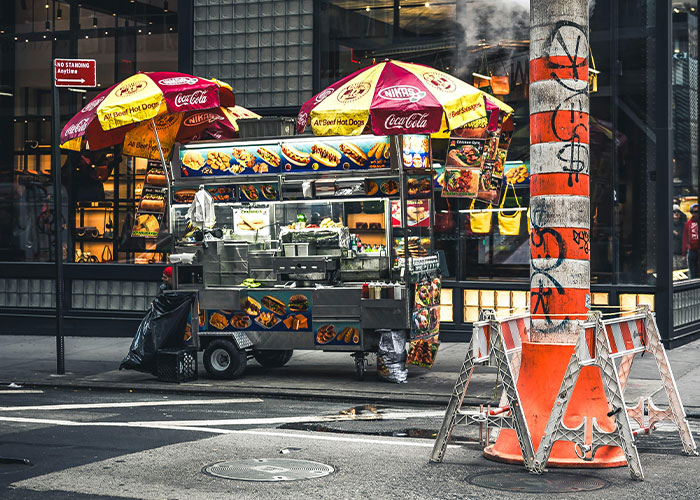 Street food cart with colorful umbrellas and smoke rising near a busy urban intersection showing chaotic scenes.