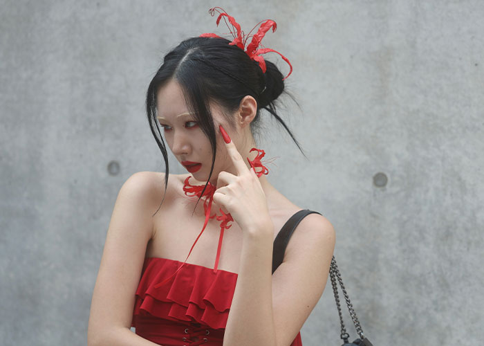 Young woman in a red dress with elaborate hair accessories and long nails posing thoughtfully against a concrete wall.