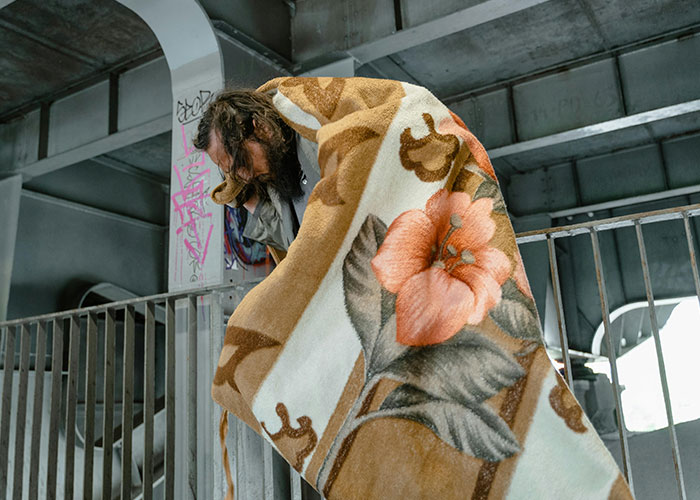 Person wrapped in a floral blanket under a bridge, portraying one of the absolutely chaotic scenes witnessed in urban settings.