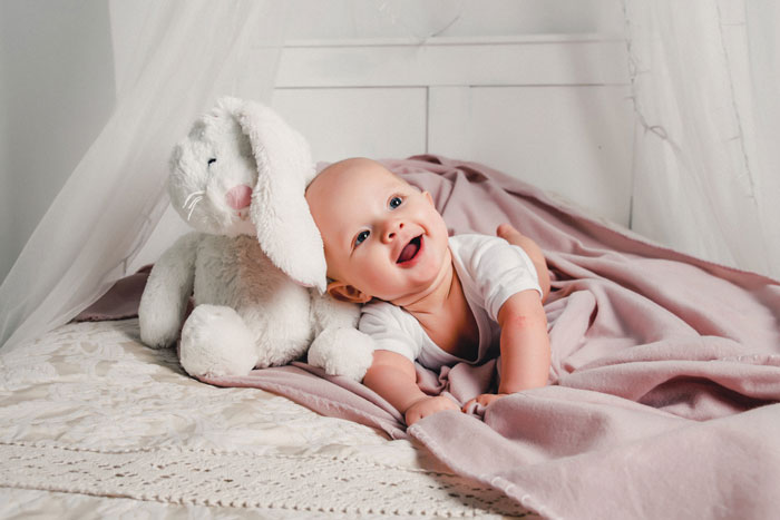 Happy baby lying on a bed under a blanket next to a plush bunny, symbolizing irresponsible teen parenting challenges. Happy baby lying on a bed under a blanket next to a plush bunny, symbolizing irresponsible teen parenting challenges.