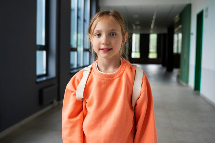 Young girl in orange sweatshirt with backpack in school hallway, representing daughter affected by native language ban conflict. Young girl in orange sweatshirt with backpack in school hallway, representing daughter affected by native language ban conflict.