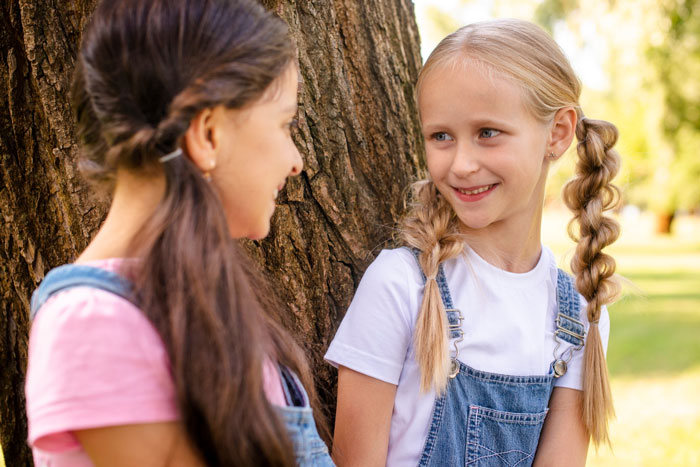 Two young girls smiling and talking near a tree, highlighting a daughter's use of her mom’s native language banned by dad. Two young girls smiling and talking near a tree, highlighting a daughter's use of her mom’s native language banned by dad.