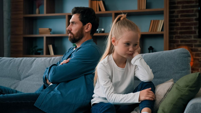 Father and daughter sitting apart on a couch looking upset, illustrating conflict over mom’s native language ban. Father and daughter sitting apart on a couch looking upset, illustrating conflict over mom’s native language ban.