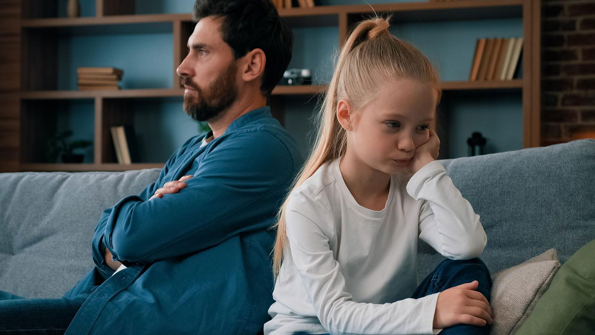Dad and daughter sitting back to back on couch after ban on using momu2019s native language causing tension.