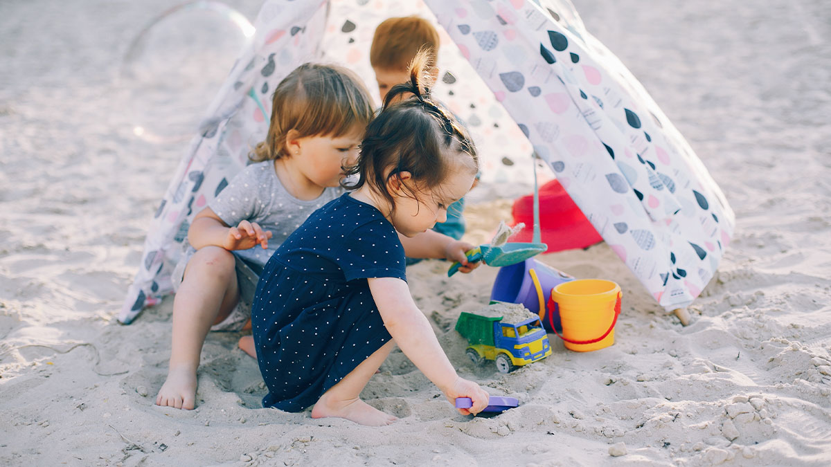 Three young children playing with sand toys under a beach tent during a family vacation outdoors.
