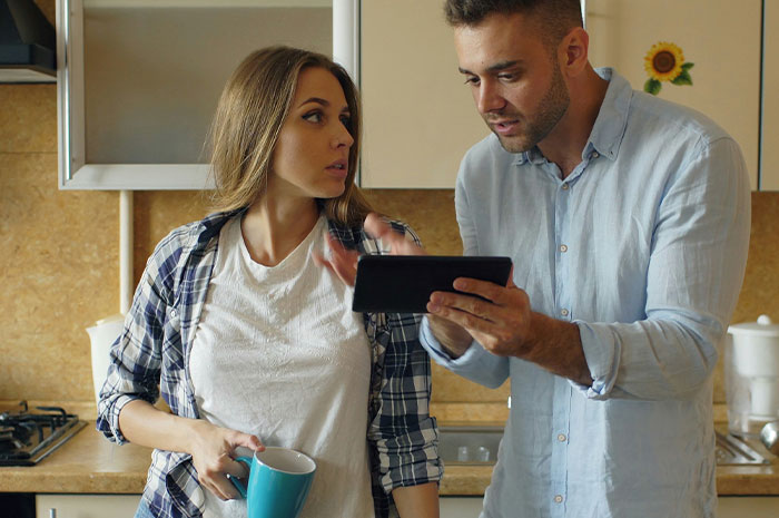 A couple discussing finances in a kitchen while looking at a tablet, highlighting choosing nose job over kids college funds. A couple discussing finances in a kitchen while looking at a tablet, highlighting choosing nose job over kids college funds.