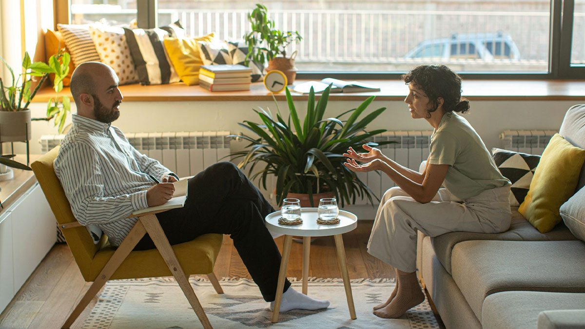Two people having a deep conversation about work secrets in a cozy, plant-filled living room setting.