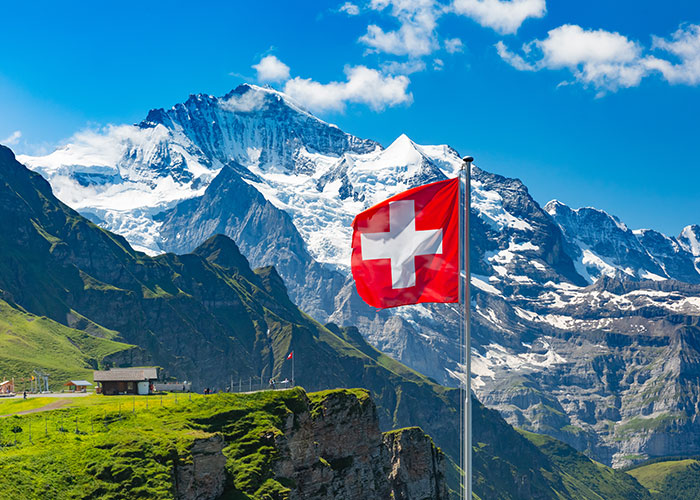 Swiss flag flying on a mountain peak with snowy Alps, illustrating the debate on whether life is better in Europe or the USA.
