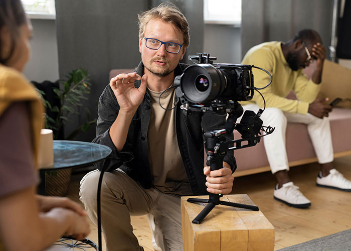 Man with glasses filming a discussion about whether life is better in the USA or Europe during a casual interview setting.