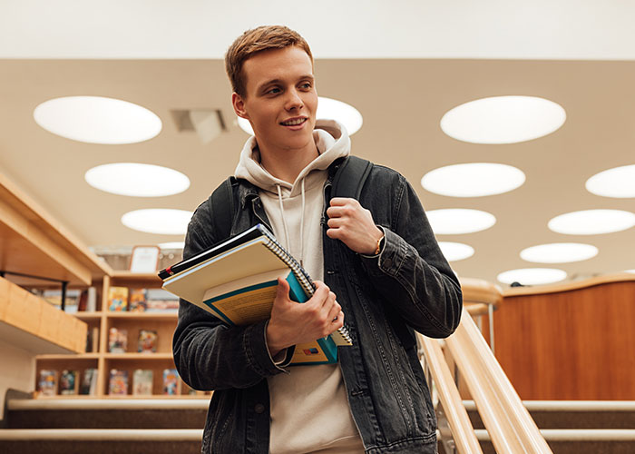 Young student holding books and backpack indoors, representing life better in the USA or Europe debate.