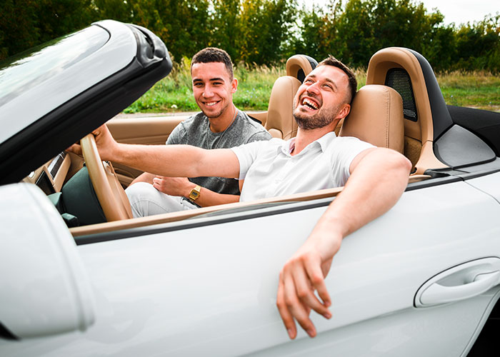 Two men laughing inside a white convertible car, enjoying life as the debate on life in the USA or Europe continues.