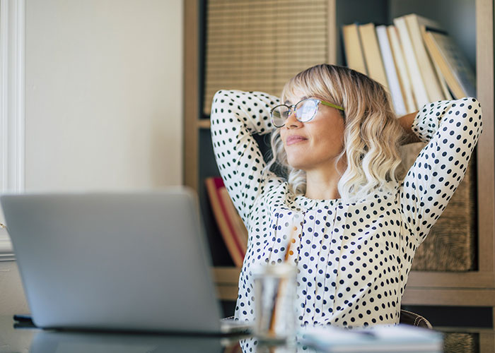 Woman with glasses relaxing at desk near laptop, reflecting on life better in USA or Europe debate.