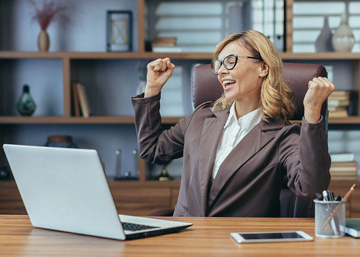 Woman celebrating at desk with laptop, expressing joy while exploring life better in USA or Europe debate online.
