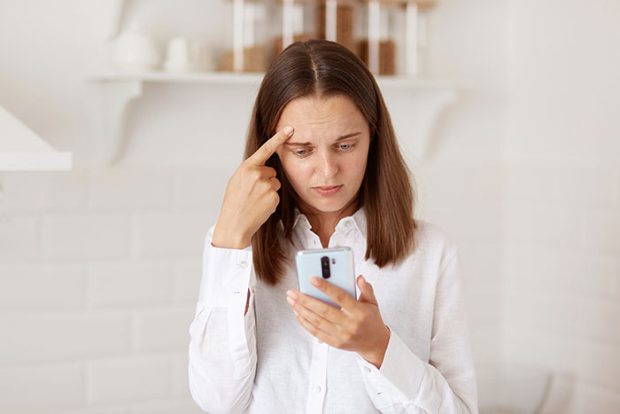 Woman in white shirt looking confused and concerned while checking phone, reflecting shock over half-sis medical bills lie. Woman in white shirt looking confused and concerned while checking phone, reflecting shock over half-sis medical bills lie.