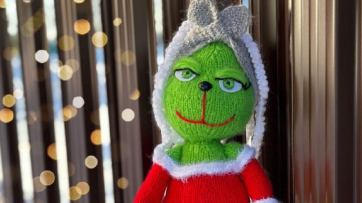 Knitted Grinch toy wearing a red and white outfit with blurred holiday lights in the background.