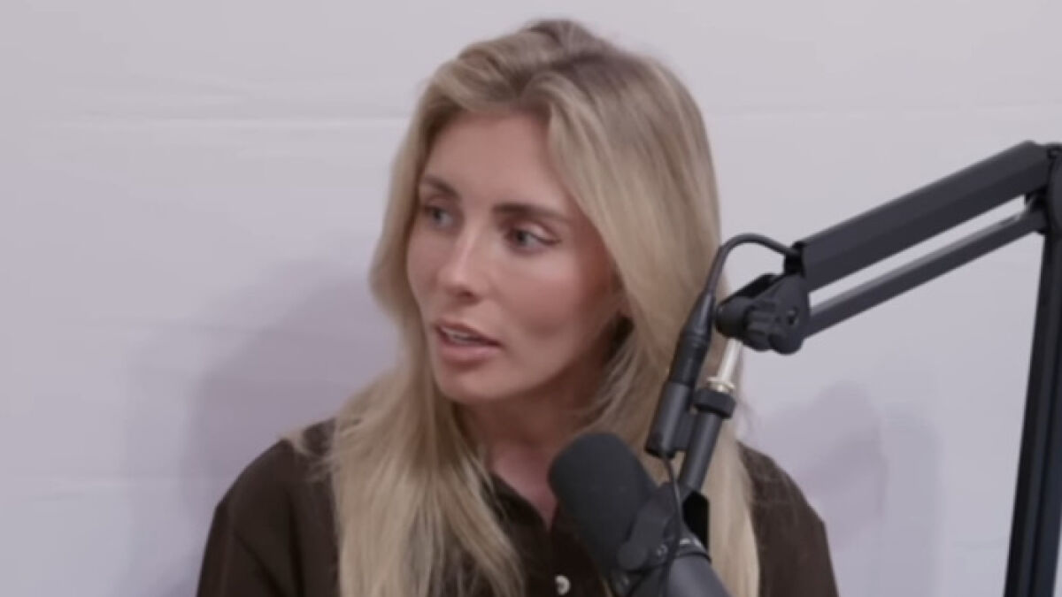 Blonde woman speaking into a microphone during a YouTuber interview with hazmat setup in the background.