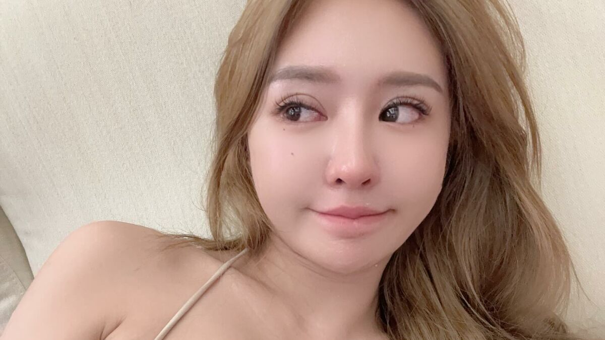 Young provocative influencer with long hair looking sideways, head resting on a light-colored couch indoors.