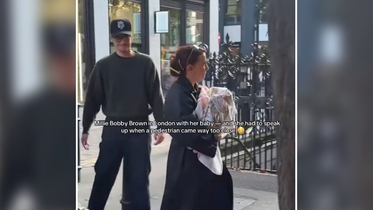 Jake Bongiovi standing behind Millie Bobby Brown as she protects their baby on a city sidewalk.