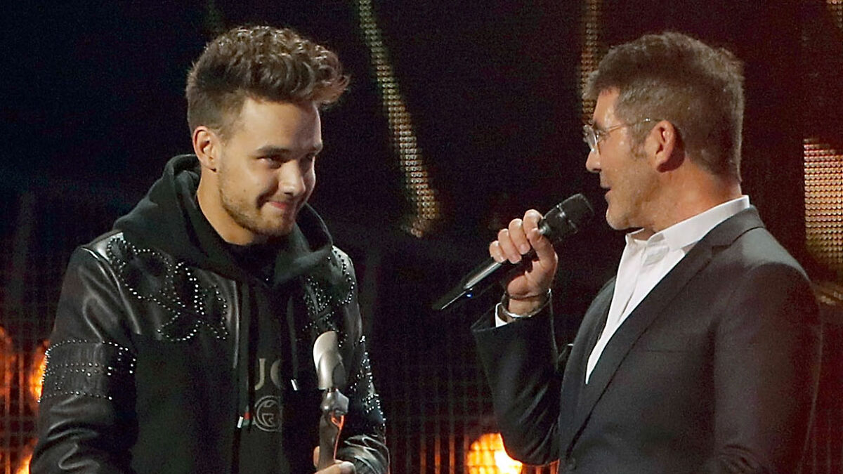 Simon Cowell speaking into a microphone while Liam Payne listens, both on a dimly lit stage.