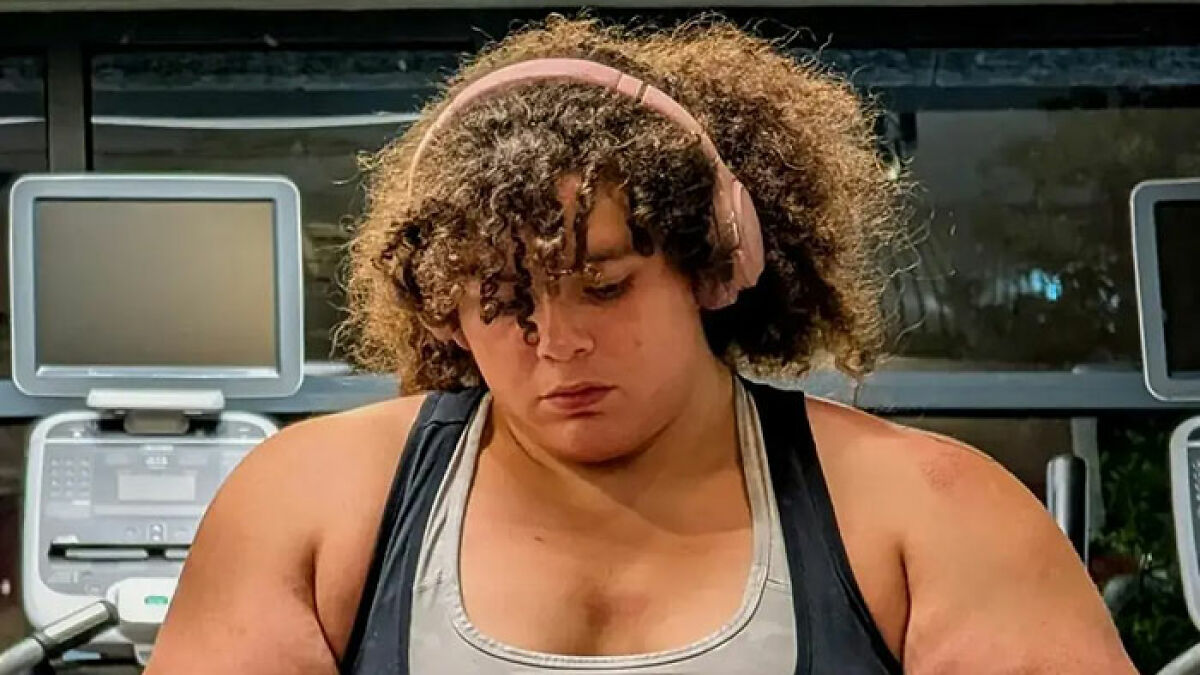 Woman with curly hair wearing headphones at gym equipment, related to worldu2019s strongest woman controversy.