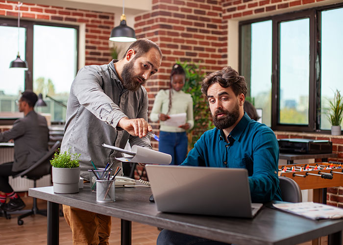 Two men discuss work at a desk with a laptop in a modern office, highlighting an expensive $1.8K office chair nearby.