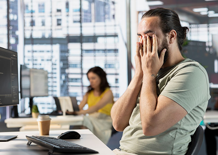 Man frustrated at office desk near computer, showing stress over expensive $1.8K office chair in a modern workspace.